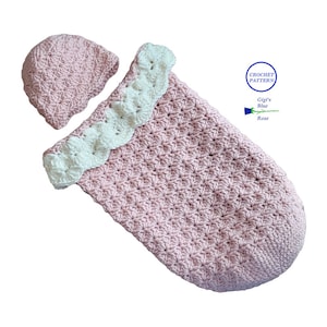 May include: A pink crocheted baby sleeping bag with a white crocheted trim and a matching pink crocheted hat. The sleeping bag has a textured pattern and the hat has a simple design. The pattern is called "Gigi's Blue Rose".