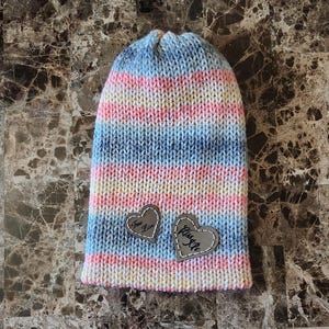 May include: A hand-knitted beanie in pastel colors of pink, blue, and yellow. The hat features two heart-shaped patches with the words "True Love" and "Kiss Me" stitched on them. The beanie is made of soft yarn.