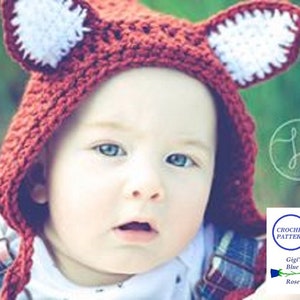 以下が含まれることがあります： 白い耳が付いた赤い毛糸の狐の帽子をかぶった赤ちゃん。帽子には顎の下で結ぶ長い尻尾が付いている。赤ちゃんは青と白のチェックのシャツを着ている。