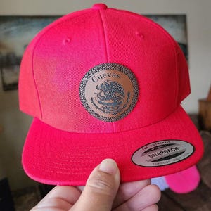 May include: A bright red snapback hat with a flat brim. The hat features a circular patch with the word "Cuevas" and an emblem. The tag reads "The Authentic Snapback Yupoong Since 1974".