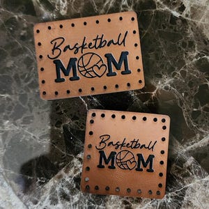May include: Two rectangular tan leather patches featuring pre-punched holes along the edges. The text "Basketball MOM" is engraved in black, with a basketball graphic replacing the letter "O" and a small heart shape integrated into the design.
