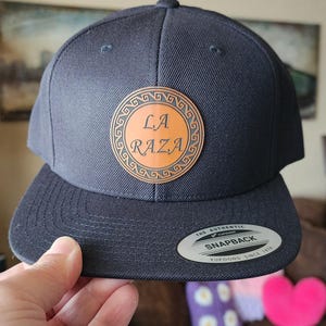 May include: A navy blue snapback hat with a flat brim. The hat features a circular, tan leather patch with the words "LA RAZA" in a stylized font. A small, silver tag is on the brim.