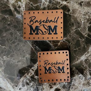 May include: Two rectangular tan leather patches featuring pre-punched holes along the edges for sewing. The patches are engraved with the text Baseball MOM in black, with the letter O replaced by a baseball graphic containing a small heart.