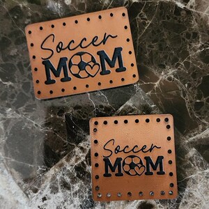 May include: Two rectangular tan leather patches featuring pre-punched holes along the edges. The text "Soccer MOM" is engraved in black, with a soccer ball replacing the letter "O" and a small heart shape integrated into the design.