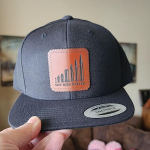 May include: A navy blue snapback hat with a flat brim. The hat features a brown leather patch with the text "SIZE DOES MATTER" and an illustration of bullets. The hat is held by a hand.