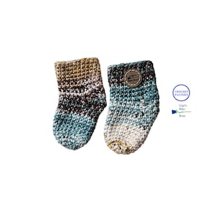 May include: A pair of crocheted baby socks in a blue, brown and beige colour scheme. The socks have a striped pattern and are made with a soft yarn.