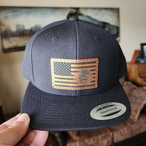 May include: A navy blue snapback hat with a flat brim. The hat features a rectangular patch with an American flag design and a gold emblem. The hat has a white and silver tag that reads "The Authentic Snapback Yupoong Since 1974".