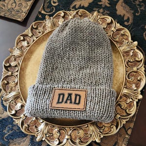 May include: A gray knit beanie with a folded brim. A brown leather patch with the word "DAD" in black lettering is sewn on the front. The hat is displayed on a gold-colored decorative plate.