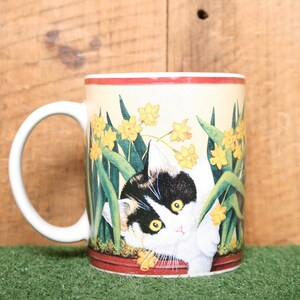 May include: White ceramic mug with a colorful illustration of a cat peeking out from a pot of yellow daffodils. The mug has a red rim and a red band around the bottom.