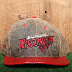 Może przedstawiać: Czapka bejsbolowa z brązowej skóry z czerwonym daszkiem. Z przodu czapki znajduje się czerwone haftowane logo z napisem "Badgers Wisconsin" z czerwoną maskotką w środku.