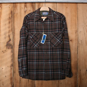 Op de afbeelding: Een bruin, zwart en rood geruit flanellen overhemd met lange mouwen en een knoopsluiting. Het overhemd heeft twee borstzakken en een label met de tekst "The Shirt".