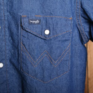 May include: A blue denim long-sleeve shirt with a pointed chest pocket and a Wrangler label. The shirt has a button-down front and a single button on each cuff.