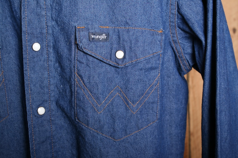May include: A blue denim long-sleeve shirt with a pointed chest pocket and a Wrangler label. The shirt has a button-down front and a single button on each cuff.
