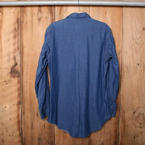 May include: A blue denim long-sleeve shirt hanging on a wooden hanger. The shirt has a pointed collar and a button-down front.