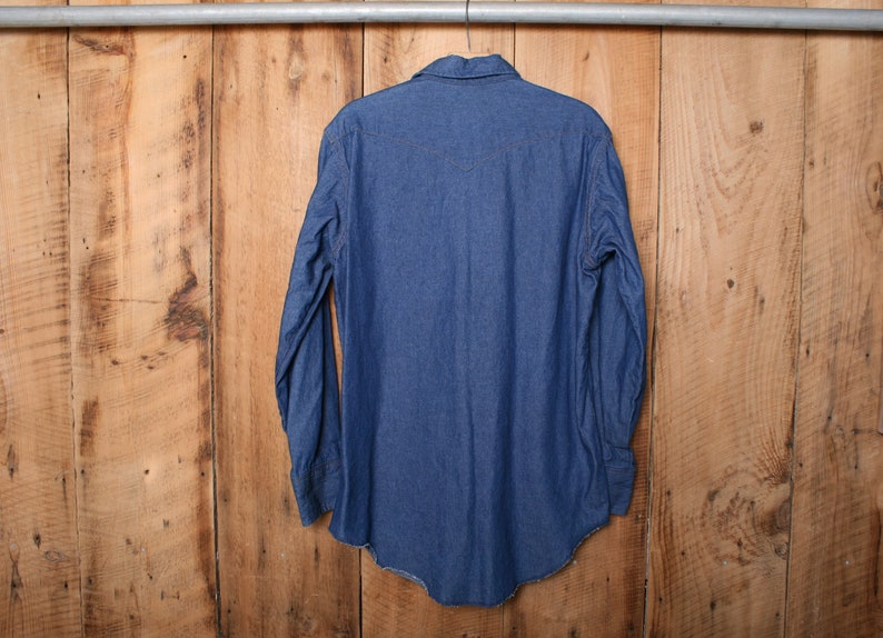 May include: A blue denim long-sleeve shirt hanging on a wooden hanger. The shirt has a pointed collar and a button-down front.