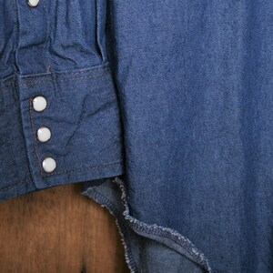 May include: Close-up of a blue denim shirt with white buttons on the cuff. The shirt is made of a soft, comfortable fabric and has a classic western style.