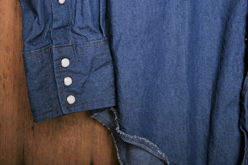 May include: Close-up of a blue denim shirt with white buttons on the cuff. The shirt is made of a soft, comfortable fabric and has a classic western style.