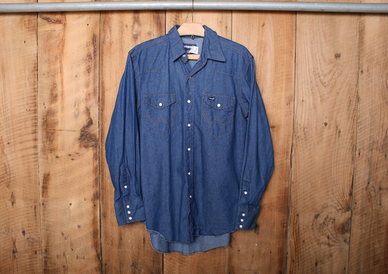 May include: A blue denim long-sleeve shirt with a button-down collar and two chest pockets. The shirt has a Western-style design with pearl snaps.