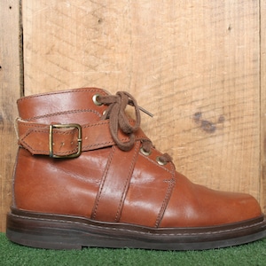 Puede incluir: Botas de tobillo de cuero marrón con una correa de hebilla y cordones. Las botas tienen una suela gruesa de goma.