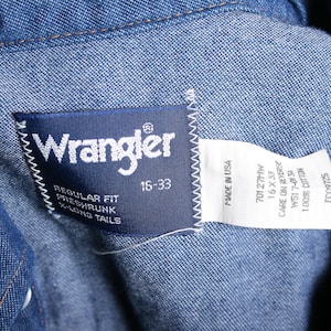 May include: A blue denim Wrangler label with white text. The label says "Wrangler", "Regular Fit", "Preshrunk", "X-Long Tails", "15-33", "Made in USA", "70127MW", "16 X 33", "Care on Reverse", "W517-0134", "100% Cotton", and "FC09725".