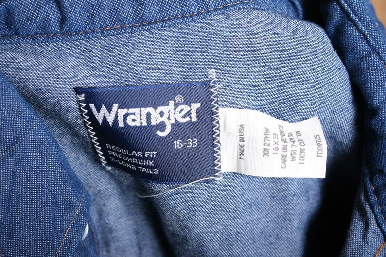May include: A blue denim Wrangler label with white text. The label says "Wrangler", "Regular Fit", "Preshrunk", "X-Long Tails", "15-33", "Made in USA", "70127MW", "16 X 33", "Care on Reverse", "W517-0134", "100% Cotton", and "FC09725".