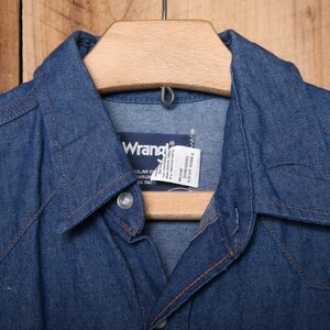 May include: A blue denim shirt with a pointed collar and a Wrangler label. The shirt is hanging on a wooden hanger.