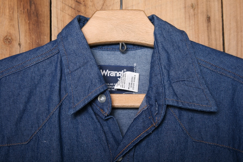 May include: A blue denim shirt with a pointed collar and a Wrangler label. The shirt is hanging on a wooden hanger.