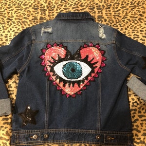May include: A dark blue denim jacket with a large pink and turquoise sequin heart with an eye design on the back. The jacket has two black star patches on the sleeves.