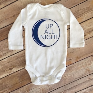 Puede incluir: Mono blanco de manga larga para bebé con un gráfico de luna creciente azul y el texto "UP ALL NIGHT".