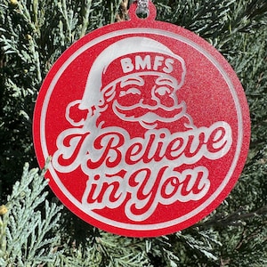 May include: Red Christmas ornament with a Santa Claus design. The ornament features the text "I Believe in You" and the initials "BMFS" on Santa's hat. The ornament is hanging from a silver string.