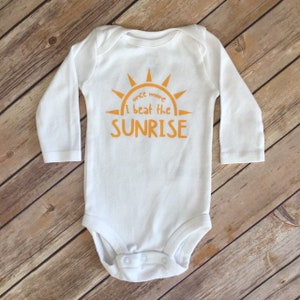 Puede incluir: Mono blanco de manga larga para bebé con un gráfico de sol amarillo y el texto "once more I beat the SUNRISE".