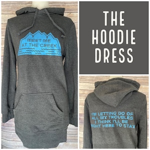 May include: A dark gray hoodie dress with a front pocket and a hood. The front features a blue graphic with the text "MEET ME AT THE CREEK". The back of the hoodie has the text "I'M LETTING GO OF ALL MY TROUBLES I THINK I'LL BE RIGHT HERE TO STAY".