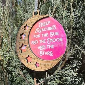 Könnte beinhalten: Ein hölzerner Halbmond-Ornament mit Sternaussparungen und einem rosa-orangenen Hintergrund. Der Ornament hat den Text "Keep Reaching for the Sun and the Moon and the Stars" in weißer Schrift. Ein silbernes Band ist zum Aufhängen befestigt.