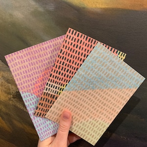 May include: A hand holds three rectangular cards with patterned designs. The cards feature a repeating dotted pattern in shades of pink, purple, and beige with black and blue accents. The background is a dark, textured surface.