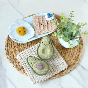Puede incluir: Juguetes de comida de ganchillo que incluyen un waffle, dos aguacates y un huevo frito. Los juguetes están dispuestos en un mantel tejido con un encaje blanco de ganchillo.