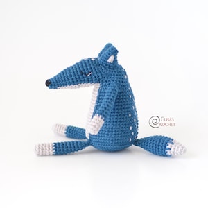 May include: A blue crocheted fox with white accents sits on a white surface. The fox has a long snout, small ears, and a long tail. The fox is a handmade toy.