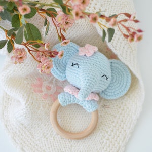 May include: A light blue crocheted elephant rattle with pink accents and a wooden ring. The rattle is lying on a white knitted blanket.