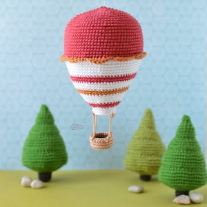 May include: A crocheted hot air balloon with red, white, and orange stripes, a brown basket, and a string hanging from the top. The balloon is hanging in front of a blue and white background with three green crocheted trees.