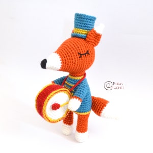 Peut inclure: Un renard en crochet portant un chapeau bleu avec un bandeau jaune, tenant un tambour rouge et jaune. Le renard porte une chemise rayée bleue et jaune.