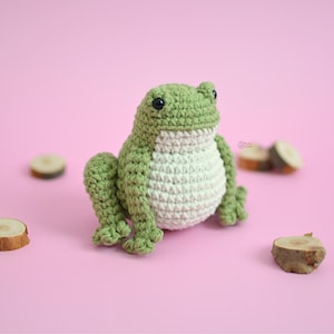 Puede incluir: Una rana de ganchillo hecha a mano en tonos verdes y blancos se encuentra sobre una superficie rosa. La rana tiene ojos negros y está rodeada de pequeñas rodajas de madera. El cuerpo de la rana es de color verde claro, con vientre blanco.