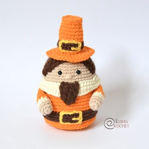 May include: A crocheted pilgrim doll wearing a brown and orange hat with a buckle. The doll has a brown beard and a white collar.