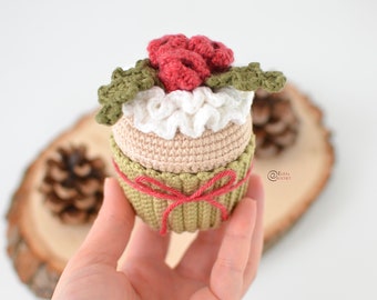May include: A crocheted cupcake with a red and white frosting and green leaves on top. The cupcake is tied with a red ribbon and is sitting on a wooden surface.
