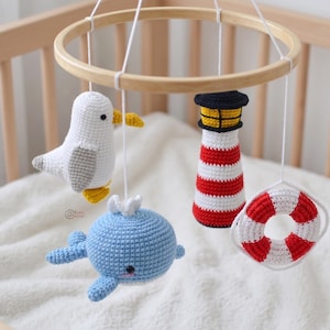 May include: A handcrafted baby mobile with a wooden ring and crocheted figures. The figures include a white seagull, a blue whale, a red and white striped lighthouse, and a red and white life preserver. The mobile is suspended over a white surface.