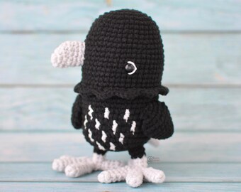 Peut inclure: Un oiseau crocheté fait à la main en noir et blanc. L'oiseau a un corps, une tête et des ailes noirs, avec des accents blancs sur le bec, les pieds et la poitrine. Il a un petit œil blanc et se tient sur une surface bleu clair.