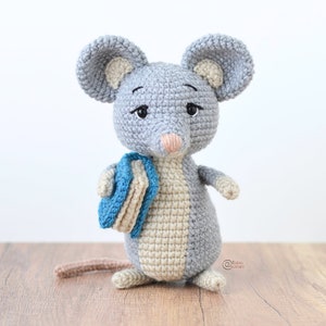 Puede incluir: Un ratón de crochet gris que sostiene una pila de libros azules y beige. El ratón tiene la nariz rosa y los ojos negros.