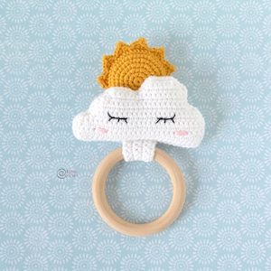 Può includere: Una nuvola all'uncinetto con un viso sorridente e un sole giallo in cima, attaccata a un anello da dentizione in legno.