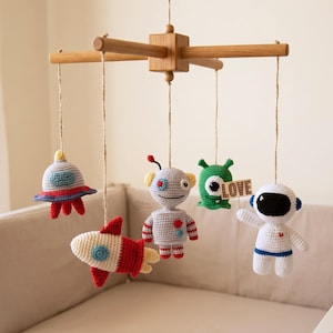 Op de afbeelding: Een handgemaakte baby mobile met gehaakte figuren met ruimtethema. De mobiel bevat een raket, ruimteschip, robot, groene alien en astronaut, allemaal opgehangen aan een houten frame. Het woord "LOVE" staat op een klein houten bord.