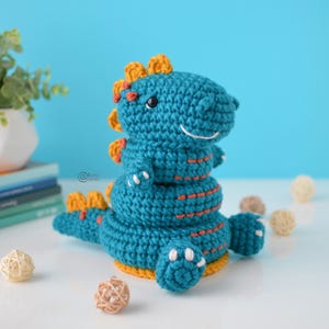May include: A teal blue crocheted dinosaur toy with orange and yellow accents. The dinosaur has a smiling face and is sitting on a white surface.