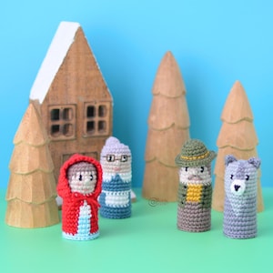 May include: Handmade crocheted finger puppets of Little Red Riding Hood, a grandmother, a hunter, and a bear, with a gingerbread house and wooden trees. The background is blue, and the puppets are made with colourful yarn.
