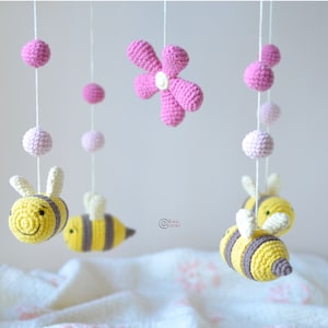 Peut inclure: Un mobile bébé en crochet rose et jaune avec des abeilles, des fleurs et des boules suspendues à un plafond blanc.